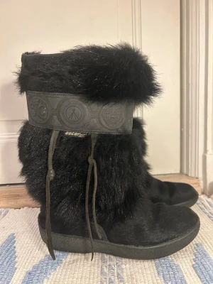 Svarta pälsboots från Oscar, stl 39 - Säljer ett par svarta boots från Oscar i storlek 39. Skorna är täckta av mjuk äkta päls och har ett brett läder dekorband med mönster upptill. De har snördetaljer på sidorna och en platt sula, perfekta för kalla dagar. Tillverkad i Italien