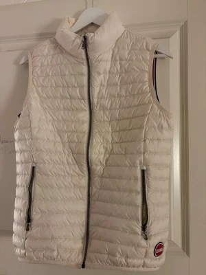 Vit dunväst från Colmar - Säljer en snygg vit dunväst från Colmar med quiltad design och hög krage. Västen har två fickor med dragkedja och en diskret logga nertill. Perfekt att slänga över en hoodie eller tröja för extra värme. Kan gå ner i pris vid snabb affär!