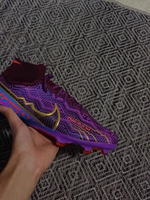 Nike fotbollsskor i lila och guld - Säljer ett par sjukt snygga Nike fotbollsskor i lila med guldiga detaljer och coola mönster. Skorna har snörning, hög strumpliknande krage och färgglad sula i blått, lila och rött. Perfekta för dig som vill sticka ut på planen!