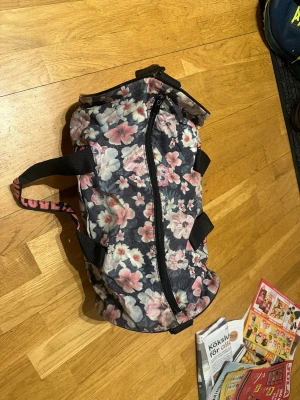 Blommig träningsväska  - Säljer en blommig träningsväska i polyester med rosa, vita och ljusblå blommor på mörk botten. Väskan har svarta handtag och axelrem samt coola rosa band med soprtig –LOVE– text. Perfekt storlek för gymmet eller träningen och stängs med dragkedja.