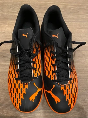 Svarta och orangea Puma fotbollsskor - Säljer ett par riktigt snygga fotbollsskor från Puma i svart och orange med coolt mönster och tydlig logga. De är i nyskick. Skorna har snörning och ovandel i syntetmaterial, perfekt för dig som vill sticka ut på planen.