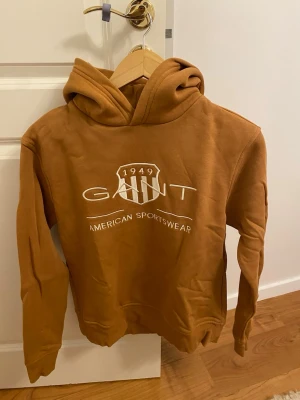 Brun hoodie från GANT  - Säljer en snygg brun hoodie från GANT logga på bröstet. Tröjan har huva och långa ärmar, perfekt för chill dagar. Klassisk och stilren design med ribbade muddar och mjukt material.