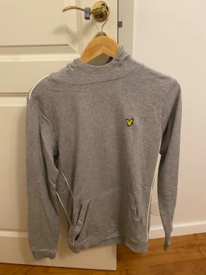 Grå hoodie från Lyle & Scott - Grå hoodie från Lyle & Scott med klassisk gul logga på bröstet. Tröjan har huva, långa ärmar och vita detaljer längs sidorna. Tillverkad i mjuk bomull, perfekt för chill dagar. Stor ficka framtill för mobilen eller händerna.