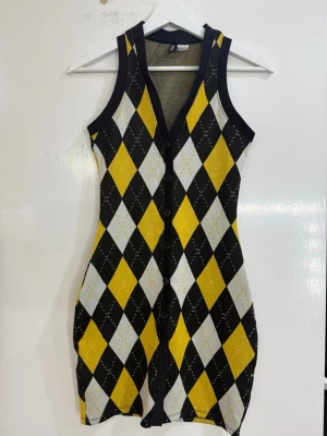 Gul och svart ärmlös kortklänning - Snygg ärmlös kortklänning med v-ringning och knappar framtill. Klänningen har ett rutigt mönster i gult, svart och vitt och är figurnära. Perfekt för dig som vill sticka ut med en cool retrostil.