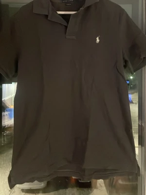 Grå pikétröja från Polo Ralph Lauren - Klassisk grå pikétröja från Polo Ralph Lauren i bomull med vit broderad logga på bröstet. Tröjan har krage och kort ärm, perfekt för en clean och stilren look. Classic fit-modell som passar till det mesta.