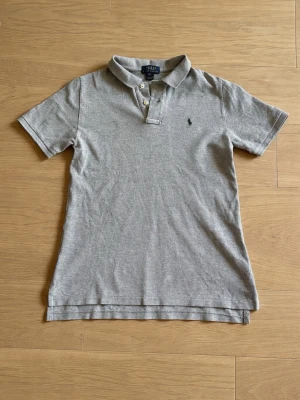 Grå pikétröja från Ralph Lauren  - Klassisk grå pikétröja från Ralph Lauren med korta ärmar och krage. Tillverkad i mjuk bomull. 