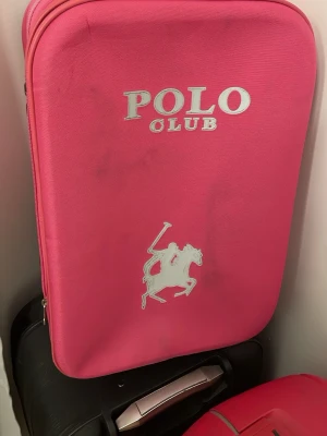 Rosa resväska från Polo Club - Säljer en snygg rosa resväska från Polo Club med stort logotryck och ikonisk polospelare på framsidan. Väskan har dragkedja runtom och är tillverkad i slitstarkt syntetmaterial. Perfekt för dig som vill resa med stil!
