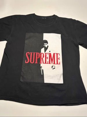 Supreme Scarface svart t-shirt - Supreme t-shirt i svart med ikoniskt Scarface-motiv på framsidan och stor röd SUPREME-text. Baksidan har ett citat från filmen tryckt i vitt. Klassisk rund hals och korta ärmar.