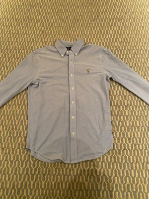 Blå skjorta från Ralph Lauren M - Säljer en klassisk ljusblå skjorta från Ralph Lauren med button-down krage och broderad logga på bröstet. Skjortan är i mjuk bomull och har långa ärmar samt knappar framtill. Perfekt till jeans eller chinos för en clean look.