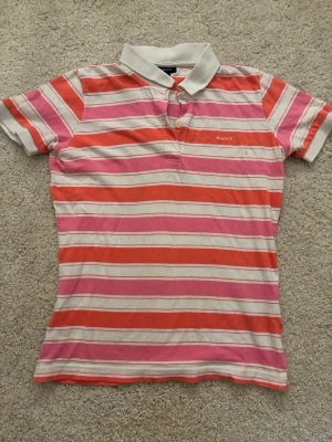 Randig skjorta från GANT i rosa och rött - Säljer en färgglad randig pikétröja från GANT i storlek   men känns som en S-M. Den har däremot några små fläckar som man inte märker. Tillverkad i mjuk bomull som känns skön mot huden. Perfekt för sommar när man ska va ute i solen.