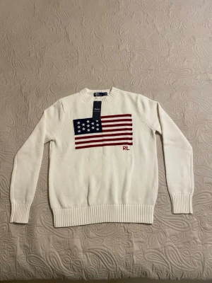 Vit stickad tröja Polo Ralph Lauren - Säljer en vit stickad tröja från Polo Ralph Lauren med amerikansk flagga och RL-brodyr framtill. Tröjan har rund halsringning, ribbade muddar och är långärmad. Perfekt för dig som gillar klassisk och stilren design.