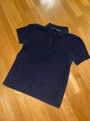 Svart pikétröja från J.Lindeberg - Säljer en marinblå pikétröja från J.Lindeberg med klassisk krage och halv dragkedja framtill. Tröjan har korta ärmar och diskret JL-logga broderad på bröstet. Tillverkad i ett mjukt bomullsmaterial som känns skönt mot huden.