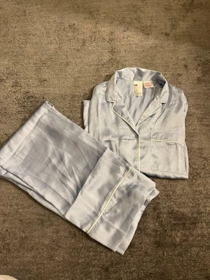 Ljusblå satinpyjamas från H&M - Säljer ett set med ljusblå pyjamas från H&M i mjuk satin. Skjortan har klassisk krage, bröstficka och vita kontrastsömmar. Byxorna har resår i midjan och samma vita detaljer vid benslut. Perfekt för mysiga kvällar hemma.