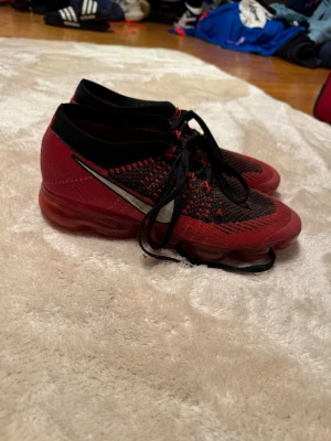 Nike Vapormax Flyknit röd/svart - Säljer ett par Nike Vapormax Flyknit sneakers i rött och svart med vit swoosh. Skorna har en stickad ovandel, svarta skosnören och den ikoniska genomskinliga röda sulan med luftbubblor. Perfekta för dig som vill ha en lätt och snygg sneaker med modern look.