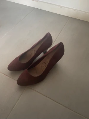 Vinröda mockapumps med låg klack - Säljer ett par vinröda pumps från DS i storlek 37. Skorna har en spetsig tå och är tillverkade i mjuk mocka med låg klack och platt sula. Snyggt minimalistisk design som ger en elegant vibe. Perfekt för dig som gillar stilrena detaljer och mörka färgtoner.