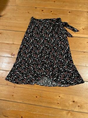 Blommig kjol från Three Sixty Five - Supersöt svart omlottkjol med blommigt mönster i vitt, lila och rött från Three Sixty Five. Kjolen har knyt i midjan och lätt volang nertill. Perfekt för dig som gillar en flowy och feminin stil🌺