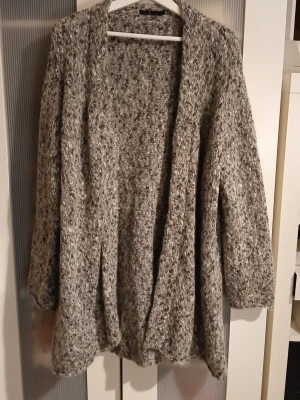 Gråmelerad kofta från Å - Mysig och oversized gråmelerad kofta från Åhléns i storlek L. Koftan är stickad med en fluffig känsla och har långa ärmar. Materialmix med mohair, ull och polyester ger en riktigt skön vibe. Perfekt att slänga på sig när det är kyligt ute.