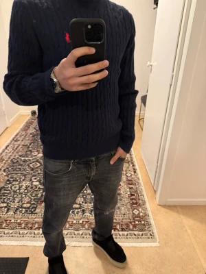 Mörkblå stickad tröja Polo Ralph Lauren - Kabelstickad mörkblå tröja Polo Ralph Lauren med klassisk rund hals och röd broderad logga på bröstet. 