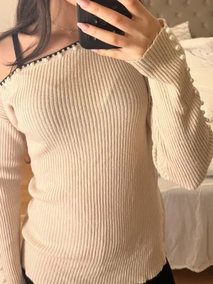 Beige ribbad offshoulder tröja med pärlor - Beige ribbad offshoulder tröja med långa ärmar. Tröjan har snygga pärldetaljer längs halsringningen och ärmsluten, samt en svart kant upptill. Okänd storlek men skulle gissa på att den är storlek S, däremot är den mycket stretchig.