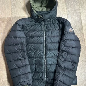 Svart Colmar pufferjacka med huva - Säljer en svart pufferjacka från Colmar med huva och dragkedja framtill. Jackan har ett quiltat mönster och är vadderad för extra värme. Perfekt för kyliga dagar och har en klassisk look med diskret logga på ärmen.