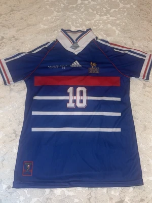 Frankrike 1998 Zidane tröja  - Retro Frankrike landslagströja från VM 1998 med Zidane och nummer 10 på ryggen. Blå med vita och röda detaljer, klassisk krage och Adidas-logga. Tillverkad i polyester med HEAT.RDY-teknologi för extra komfort. Perfekt för fotbollsfans!