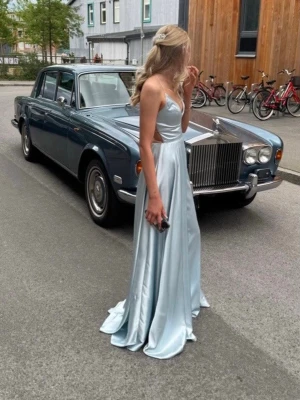 Ljusblå balklänningen öppen rygg med snörning  - Säljer en ljusblå balklänning i satin med v-ringad front och öppen rygg med snygg snörning. Klänningen har en hög slits som syns när man går. Använd en gång köpt från ivory field. Är lite smutsig längst ner så kan behöva tvättas. 