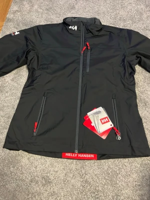 Svart vindjacka från Helly Hansen - Säljer en svart vindjacka från Helly Hansen med röda detaljer och dragkedjor. Jackan har flera fickor med dragkedja och en mjuk fleecefodrad insida för extra komfort. Perfekt för blåsiga dagar och har en sportig look med HH-logga på ärmen jackan är helt ny 