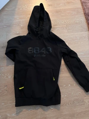Svart skid/vindjacka från 8843 Altitude - Säljer en svart vindjacka med huva från 8843 Altitude. Jackan har stor logga framtill, dragsko i huvan och gula detaljer vid fickorna. Perfekt för blåsiga dagar och enkel att matcha med det mesta.