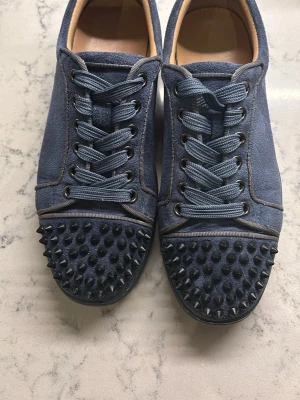 Blå mockasneakers från Christian Louboutin - Säljer ett par blå sneakers i mocka från Christian Louboutin med ikonisk röd sula och coola svarta nitar på tån. Skorna har snörning och en rund tå, vilket ger en edgy vibe till din outfit. Perfekta för dig som vill sticka ut med exklusiva detaljer.