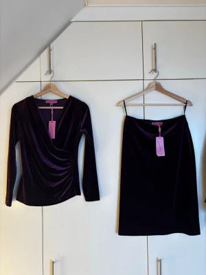 Nya oanvända lila sammetstop och kjol från Camilla Thulin storlek 38 - Supersnygg lila sammettop och kjol från Camilla Thulin. Topen har omlottliv med v-ringning, långa ärmar. Kjolen är rak. Mjuk, glansig finish som ger en lyxig känsla. Nya oanvända!