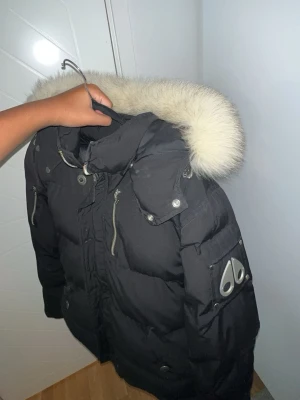 Svart dunjacka med päls från Moose Knuckles - Säljer en svart dunjacka från Moose Knuckles med stor fluffig päls runt huvan och coola metalldetaljer. Jackan har dragkedjor, knappar och logga på ärmen. Perfekt för kalla dagar och riktigt snygg streetstil.