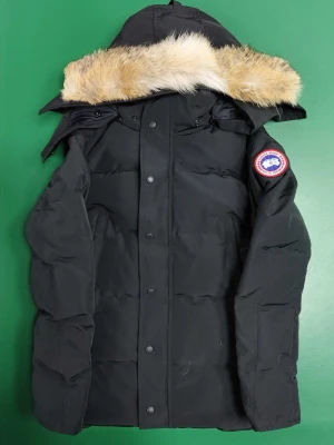 Svart dunjacka från Canada Goose XS - Säljer en svart dunjacka från Canada Goose i storlek XS. Jackan har en fluffig huva med beige pälsdetalj, klassisk logga på ärmen och stängs med både dragkedja och knappar. Perfekt för kalla dagar och riktigt snygg streetstil.