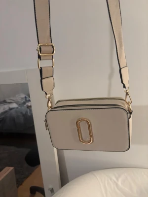 Beige axelväska  - Säljer en beige axelväska med bred justerbar axelrem och guldfärgade detaljer. Väskan har två stora fack med dragkedja och en ficka på baksidan. Klassisk rektangulär form och stilren design med logga i metall framtill.