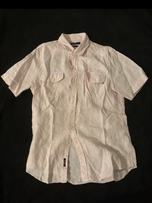 Ljusrosa linneskjorta från Replay - Snygg ljusrosa skjorta från Replay i luftig linnemix. Kortärmad modell med klassisk krage, två bröstfickor med knapp och knäppning framtill. Perfekt för varma dagar och chill stil. Replay-logga på ena fickan och vid nederkant.