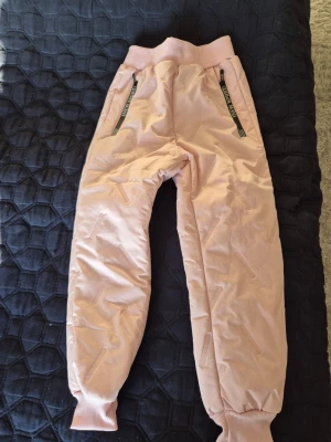 Ljusrosa täckbyxor med dragkedjor - Täckbyxor i ljusrosa med ribbad midja och muddar vid benslut. Utrustade med svarta dragkedjor framtill med texten 'NEW SPORT'. Insidan är fodrad med mjuk teddy för extra värme. Perfekta för kalla dagar och vinteraktiviteter.