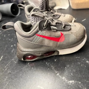 Baby Grå Nike sneakers med röd swoosh - Säljer ett par grå Nike sneakers med tydlig röd swoosh på sidan och synlig Air Max-dämpning i sulan. Ovandelen är i mesh och syntet med coola sömdetaljer och snörning. Sulan har mönstrad undersida för bra grepp. Perfekta för dig som gillar sportig stil.