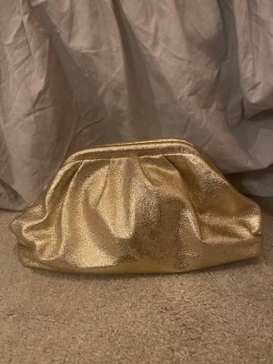 Guldig kuvertväska i syntet - Säljer en glammig kuvertväska i metallic guld med mjuk, puffig form och enkel öppning. Väskan har ett stort huvudfack med ljus insida och en liten innerficka. Perfekt för att lyfta vilken outfit som helst med extra bling.