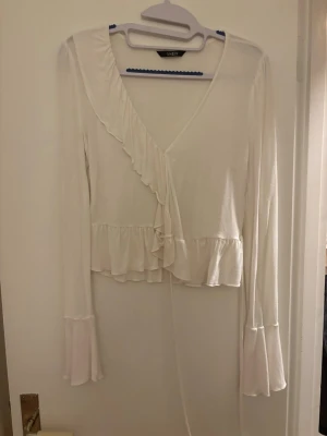 Vit omlottopp med volang - Säljer en vit omlottopp från SHEIN med långa ärmar och volangdetaljer längs kanten och nederdelen. Toppen har v-ringning och knytband. 