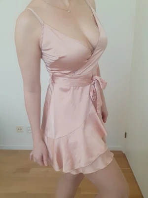 Ljusrosa omlottklänning i satin - Supersöt ljusrosa kortklänning i glansigt satinliknande material. Klänningen har omlottmodell med v-ringning, smala axelband och ett knytband i midjan. Volangdetaljer längst ner på kjolen ger en extra feminin touch.