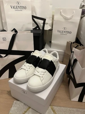 Valentino Garavani Open sneakers vit/svart - Säljer ett par Valentino Garavani Open sneakers i vitt skinn med bred svart läderdetalj över vristen. Klassisk rund tå, vita snören och diskret logga på plösen. Snygg och stilren design med platt sula och nitar på hälen för extra edge.