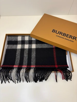 Burberry scarf - Elegant Burberry-halsduk i klassisk design och mjukt, värmande material. Tidlöst rutmönster i mörka toner med stilrena röda detaljer. Levereras i originalförpackning – perfekt både för egen stil och som exklusiv present.