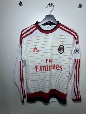 Vit AC Milan långärmad matchtröja - AC Milan långärmad matchtröja från Adidas i vitt med tunna gula ränder, röda detaljer och klubbmärke på bröstet. Tre röda ränder längs ärmarna, Champions League-märke på ärmen och Fly Emirates-tryck på magen. Tillverkad i polyester och har svarta och röda detaljer vid krage och nederkant.