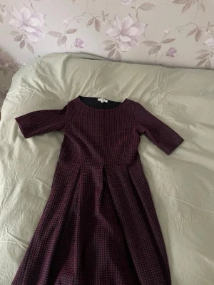Mörk rutig klänning med veck - Säljer en mörk rutig klänning med vinröda och svarta toner. Klänningen har trekvartsärm, rund halsringning och markerad midja med snygga veck i kjolen. Tyget är lite tjockare och känns strukturerat, perfekt för dig som gillar en klassisk men cool look.