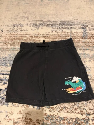 Svarta shorts med dinosaurie från H&M - Svarta shorts från H&M i mjuk bomull med elastisk midja och snörning. På ena benet finns ett färgglatt tryck av en dinosaurie som surfar med texten 'Happiness comes in waves'. Perfekta för chill dagar eller strandhäng.