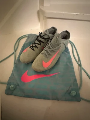 Nike Mercurial AG Elite Prism Pack - Säljer ett par Nike Mercurial Elite fotbollsskor i ljusblått med stora rosa swoosh-loggor och coola detaljer på hälen. Skorna har snörning, en platt sula med konstgräs dobbar och är tillverkade i gripknit. Ena skon har svarta skosnören och den andra ljusblåa. Perfekta för dig som vill sticka ut på planen!
