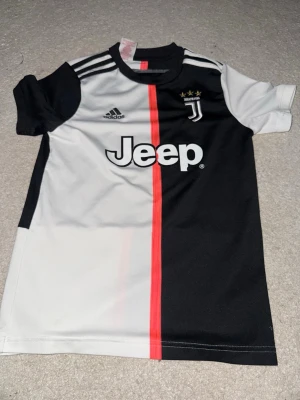 Juventus Adidas matchtröja svart/vit M - Juventus matchtröja från Adidas i svart och vitt med röd rand i mitten. Tröjan har klassiska Adidas-ränder på axlarna, Jeep-logga på bröstet och Juventus klubbmärke med tre stjärnor. Tillverkad i lätt och ventilerande polyester, perfekt för fotboll.