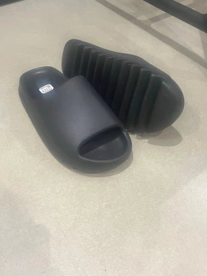 Svarta Adidas slides med räfflad sula - Säljer ett par svarta Adidas slides med chunky, räfflad sula och öppen tå. Tillverkade i syntetmaterial för en mjuk och bekväm känsla. Perfekta för sommaren och chill dagar. Diskret Adidas-logga på fotbädden.