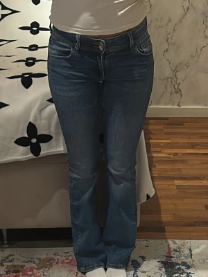 Blå bootcut jeans med fickdetalj - Säljer ett par klassiska blå jeans med bootcut-modell. Jeansen har en snygg ficka med knapp bak och sitter tajt över låren för att sedan bli vidare nedtill. Perfekta för dig som gillar retrovibbar och vill ha ett par jeans som sticker ut lite extra.