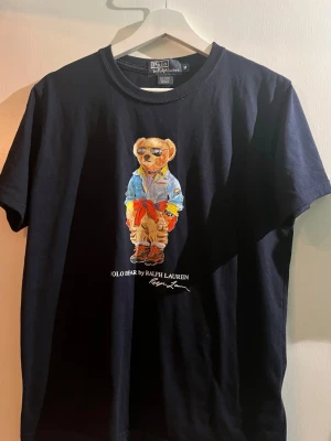 Svart Polo Bear t-shirt från Ralph Lauren - Marin blå t-shirt från Polo Ralph Lauren med ikoniskt Polo Bear-tryck på bröstet. T-shirten är i bomull och har en klassisk passform med korta ärmar och rund hals. Perfekt statement-plagg för dig som gillar streetstyle och snygga prints. Lite liten i storleken!