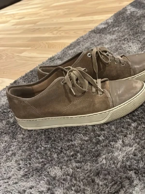 Beige sneakers från Lanvin i mocka - Säljer ett par snygga beige sneakers från Lanvin med ovandel i mocka och tåparti i skinn. Skorna har vita gummisulor och klassisk snörning. Perfekta för dig som gillar stilrena och neutrala färger. Riktigt schyssta till jeans eller chinos.